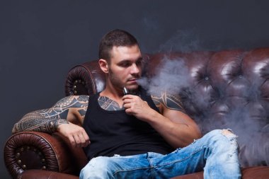 Vaper. Dövmeli adam koyu arka plan üzerinde elektronik sigara duman deri kanepede oturuyor.