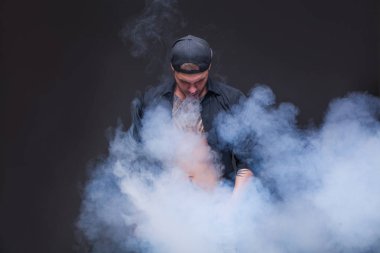 Vaper. Adam mavi kot pantolon giymiş, siyah gömlek ve siyah beyzbol şapkası dövmeler ile koyu arka plan üzerinde elektronik sigara duman