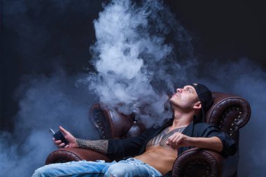 Vaper. Dövmeli adam koyu arka plan üzerinde elektronik sigara duman deri kanepede oturuyor.