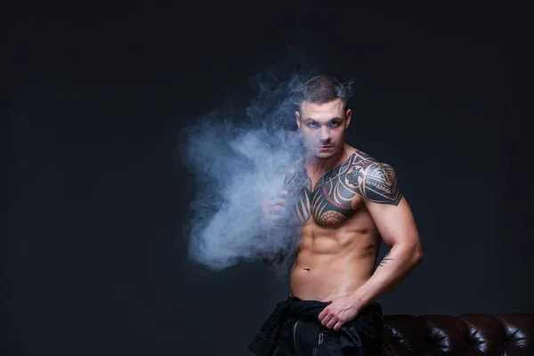 Vaper. El hombre con un torso desnudo muscular con tatuajes fuma un cigarrillo electrónico en el fondo oscuro —  Fotos de Stock