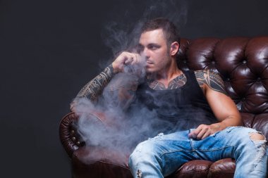 Vaper. Dövmeli adam koyu arka plan üzerinde elektronik sigara duman deri kanepede oturuyor.