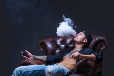 Vaper. Dövmeli adam koyu arka plan üzerinde elektronik sigara duman deri kanepede oturuyor.