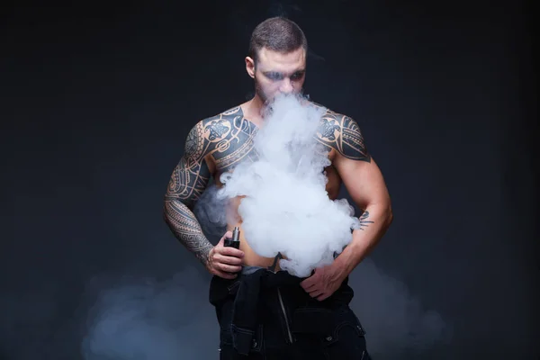 Vaper. Mannen med en muskulös nakna torso med tatueringar röka en elektronisk cigarett på den mörka bakgrunden — Stockfoto