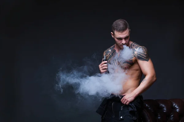 Vaper. El hombre con un torso desnudo muscular con tatuajes fuma un cigarrillo electrónico en el fondo oscuro —  Fotos de Stock