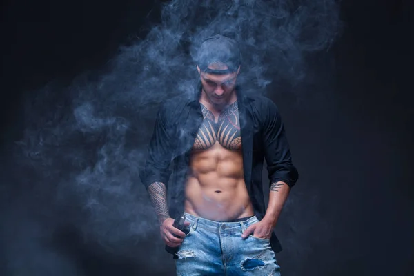 Vaper. El hombre vestido con jeans azules, camisa negra y gorra de béisbol negra con tatuajes fuma un cigarrillo electrónico sobre el fondo oscuro —  Fotos de Stock