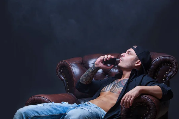 Vaper. El hombre con tatuajes se sienta en un sofá de cuero fumar un cigarrillo electrónico en el fondo oscuro . — Foto de Stock