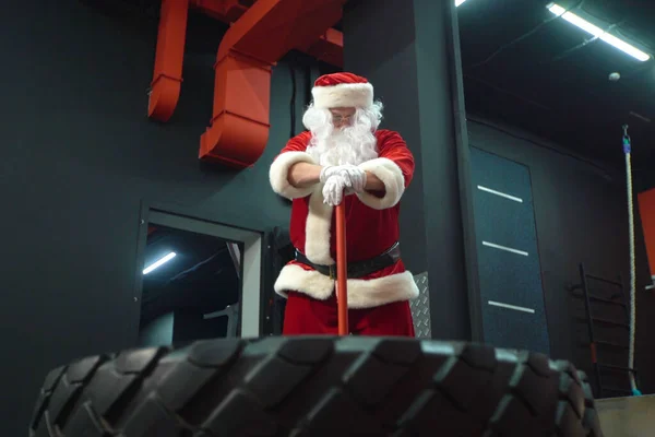 Trening Świętego Mikołaja na siłowni w Boże Narodzenie. Santa Claus uderzenie koła opony z młotkiem. — Zdjęcie stockowe