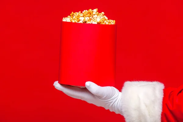 Natale. Foto di Babbo Natale mano guantata Con un secchio rosso con popcorn, su uno sfondo rosso — Foto Stock