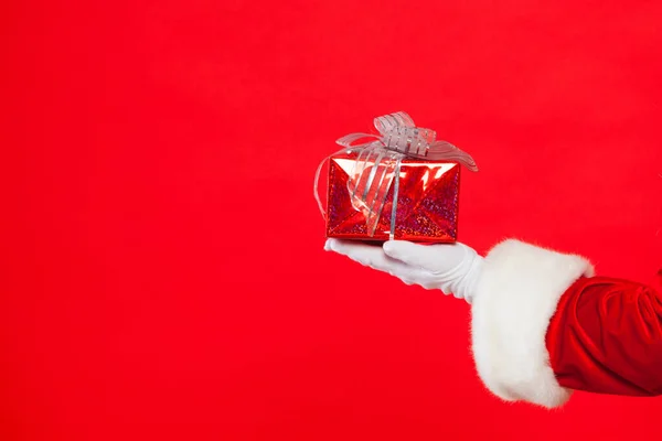 Foto der handschuhten Weihnachtsmann-Hand mit Geschenkbox, auf rotem Hintergrund. Weihnachten — Stockfoto