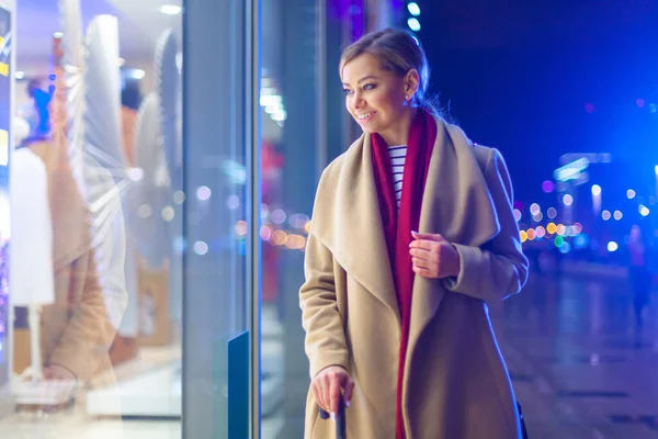 Weihnachten. eine junge schöne Frau am späten Abend blickt auf das leuchtende Fenster und sucht sich ein Geschenk aus. bunte, magische Beleuchtung, schöne Blendung. fröhliches Mädchen wunderte sich am Fenster, die Qual der — Stockfoto