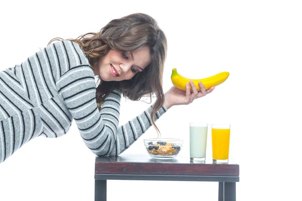 若い妊娠中の女性は、バナナがあるテーブルに座って、シリアルとプレート、ケフィアとジュースとグラスがあります。妊娠中の栄養の概念。白い背景に隔離. — ストック写真