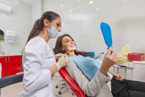 Zahnarzt. eine Frau in der Zahnarztpraxis untersucht ihre Zähne im Spiegel und lächelt. — Stockfoto