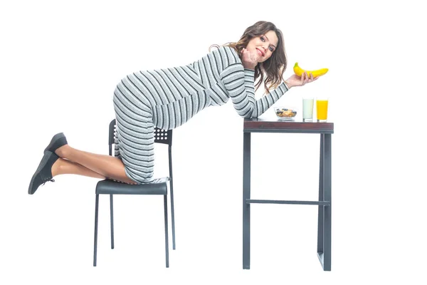 Молода вагітна жінка сидить за столом, на якому лежить банан, є тарілка з кашею, склянка з кефіром і соком. Концепція харчування під час вагітності. Ізольовані на білому тлі . — стокове фото