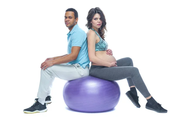 Een jong stel en hun vrouw in de laatste maanden van de zwangerschap zitten op een sportmat in de buurt van de bal. Zwarte man en blanke vrouw. Het concept van een gelukkige paar verschillende races samen bereiden — Stockfoto