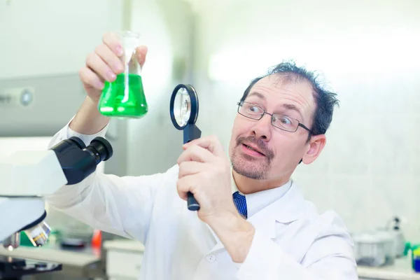 Chemik oszalał. Szalony naukowiec przeprowadza eksperymenty w laboratorium naukowym. Z zaskoczonymi oczami patrzy przez lupę na zielony płyn w kolbie.. — Zdjęcie stockowe