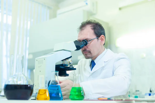 Químico loco. Un científico loco realiza experimentos en un laboratorio científico. Realiza investigaciones utilizando un microscopio . —  Fotos de Stock