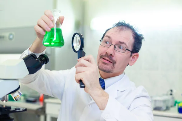 Хімік божевільний. Божевільний вчений проводить експерименти в науковій лабораторії. З здивованими очима, він дивиться через збільшувальний стакан на зелену рідину в колбі . — стокове фото