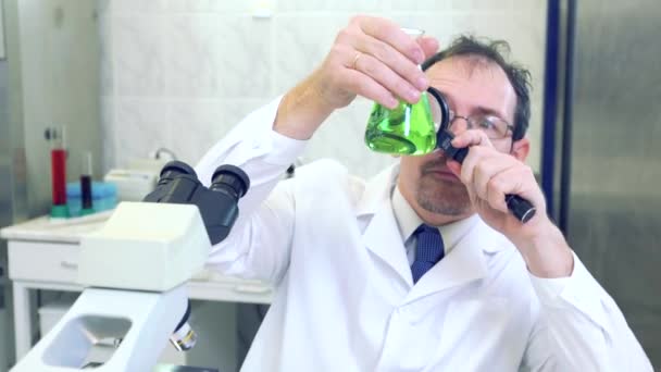 Chimiste fou. Un scientifique fou mène des expériences dans un laboratoire scientifique. Avec des yeux surpris, il regarde à travers une loupe le liquide vert dans la fiole . — Video