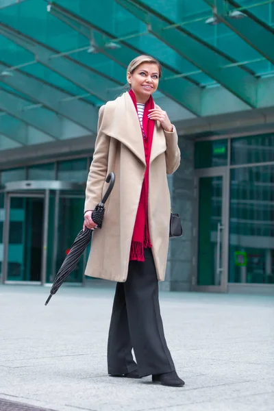 stock image Outdoors lifestyle fashion portrait of stunning brunette girl. Walking on the city street. Going shopping. Wearing stylish white fitted coat, red neckscarf, black umbrella cane. Business woman.