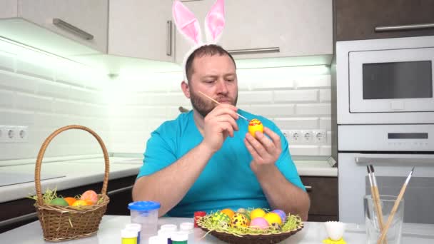 Vrolijk Pasen. Een man in een konijnenoren blauw t-shirt in de keuken schildert eieren voor Pasen. — Stockvideo