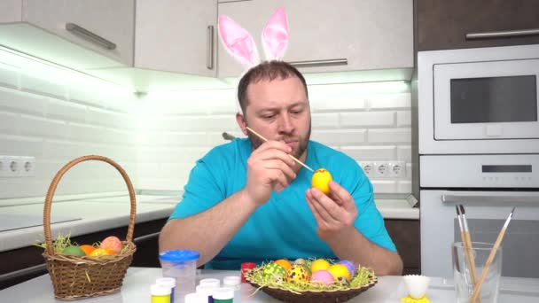 Feliz Páscoa. Um homem em orelhas de coelho camiseta azul na cozinha pinta ovos se preparando para a Páscoa . — Vídeo de Stock