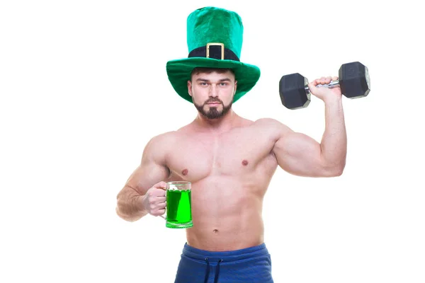Día de San Patricio. Un hombre musculoso con un sombrero verde sostiene una taza de cerveza, cerveza y una mancuerna negra. Fondo blanco aislado. ABS . — Foto de Stock