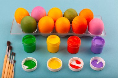 Renkli Paskalya yumurtaları ve püsküllü çok renkli guaj kavanozları şeffaf bir kutunun üst görüntüsü. Mavi arka planda izole edilmiş. Paskalya için hazırlanılıyor.