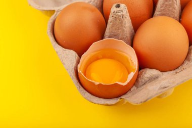 Yumurtalı tavuk yumurtası. Sarı arka planda kahverengi yumurtaları olan açık gri bir kutunun üst görüntüsü. Bir yumurtanın yarısı kırık, sarı yuvarlak sarısı. Sağlıklı yaşam tarzı kavramı, saf protein almak.