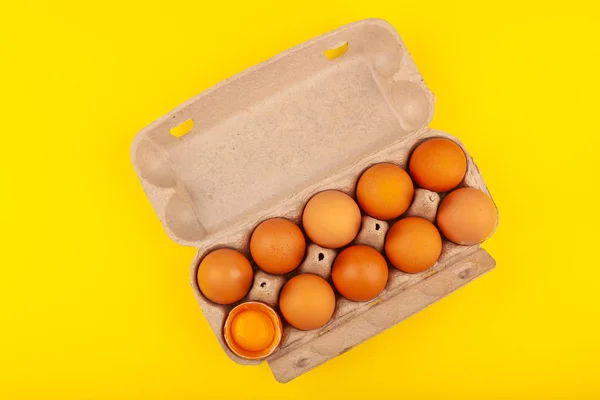 अंडी चिकन अंडी. पिवळ्या पार्श्वभूमीवर पृथक् त तपकिरी अंडी असलेल्या खुल्या राखाडी बॉक्सचे शीर्ष दृश्य. एक अंडी अर्धा तुटलेली, पिवळी गोल यॉर्क आहे. निरोगी जीवनशैलीची संकल्पना, शुद्ध प्रथिने मिळविणे — स्टॉक फोटो, इमेज