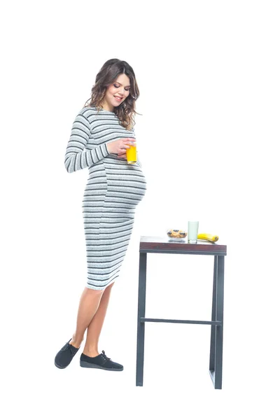 Una joven embarazada se sienta en una mesa en la que se encuentra un plátano, hay un plato con cereales, un vaso con kéfir y jugo. El concepto de nutrición durante el embarazo. Aislado sobre un fondo blanco . —  Fotos de Stock