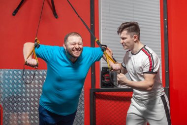 Fat man in a gym is diligently doing pull-ups under the guidance of a personal trainer. Overweight TRX