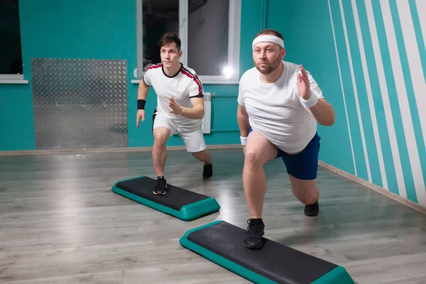 Tłuścioch bardzo ciężko pracuje wykonując ćwiczenia step-up na krokach podczas grupowych zajęć fitness. Nadwaga — Zdjęcie stockowe