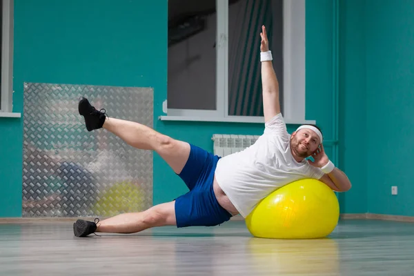 Hombre gordo sonriente está haciendo ejercicios usando la pelota de fitness. Hombre con sobrepeso está contento con el resultado de su entrenamiento en clases de fitness en grupo — Foto de Stock