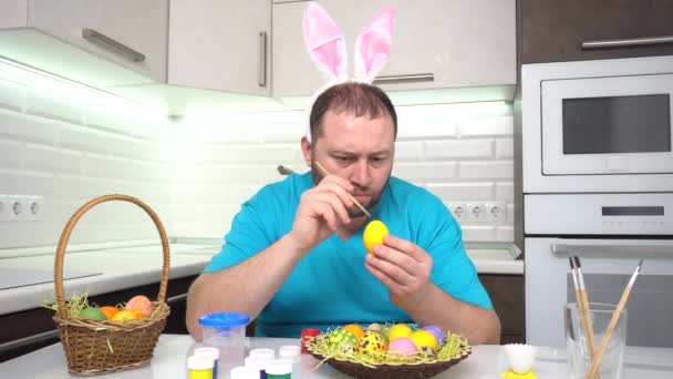Veselé Velikonoce. Muž v králičích uších, modré tričko v kuchyni, maluje vajíčka na Velikonoce.. — Stock video