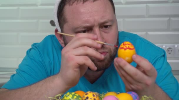 Feliz Pascua. Un hombre con una camiseta azul de orejas de conejo en la cocina pinta huevos preparándose para la Pascua . — Vídeo de stock