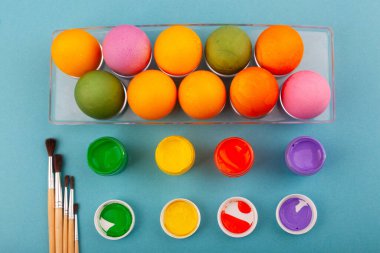 Renkli Paskalya yumurtaları ve püsküllü çok renkli guaj kavanozları şeffaf bir kutunun üst görüntüsü. Mavi arka planda izole edilmiş. Paskalya için hazırlanılıyor.