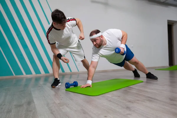 Hombre gordo está trabajando duro en el gimnasio con su entrenador personal haciendo ejercicios con pesas en el límite de las posibilidades . — Foto de Stock