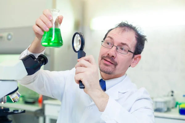 Хімік божевільний. Божевільний вчений проводить експерименти в науковій лабораторії. З здивованими очима, він дивиться через збільшувальний стакан на зелену рідину в колбі . — стокове фото