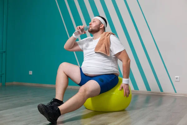 Homem com excesso de peso está sentado em uma bola de fitness exausto após um treinamento duro em aulas de fitness em grupo. Gordo bebe água com uma toalha no ombro. — Fotografia de Stock
