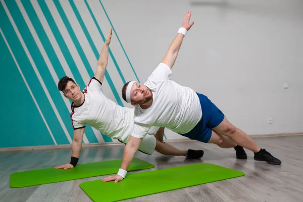 Tłuścioch ciężko pracuje na siłowni ze swoim osobistym trenerem wykonując ćwiczenia na macie na granicy możliwości — Zdjęcie stockowe