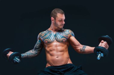 Dövmeli güçlü kaslı atletik adam siyah arka planda dambılları kaldırıyor.