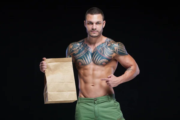 Gespierde man bodybuilder met tatoeages. Man poserend op een zwarte achtergrond met papieren zak. Het concept van reclame voor de juiste sportvoeding. Logo kopieerpasta. — Stockfoto
