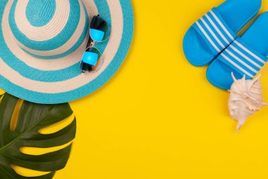 Sarı arka plandaki mavi plaj aksesuarları. Güneş gözlükleri, parmak arası terlikler, çizgili şapka, kabuk ve canavar yaprağı. Plaj tatili kavramı, deniz turu, sıcak güneşli yaz. Reklam alanı