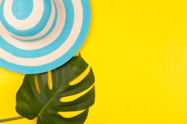 Sarı arkaplanda plaj aksesuarları, çizgili mavi şapka ve canavar yaprağı. Uzun zamandır beklenen deniz ve seyahat tatili kavramı. Reklam alanı