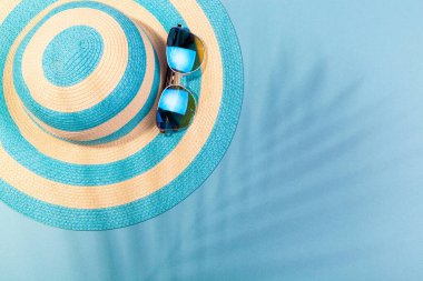 Turkuaz mavi arka planda plaj aksesuarları. Güneş gözlükleri ve palmiye yaprağının gölgesinde çizgili mavi şapka. Plaj tatili kavramı, deniz turu, sıcak güneşli yaz. Reklam alanı