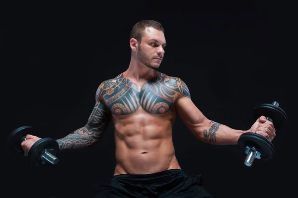 Татуйований сильний м'язистий спортсмен піднімає гантелі на чорному тлі — стокове фото
