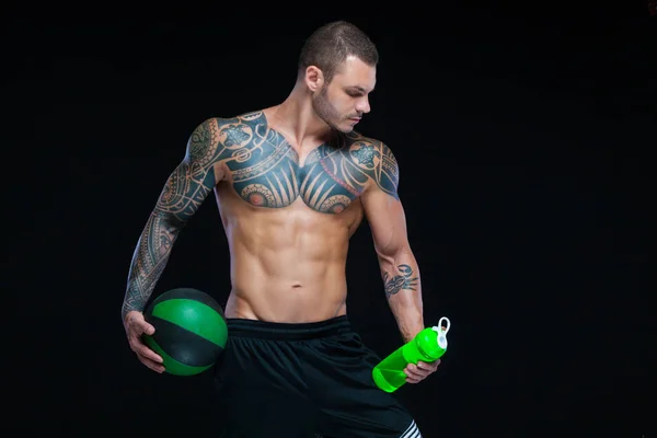 Muscular hombre culturista con tatuajes. Hombre posando sobre un fondo negro con botella de agua y bola — Foto de Stock