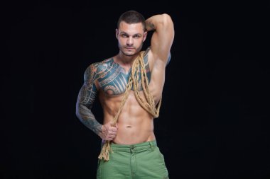 Dövmeleri olan kaslı bir vücut geliştirici. Siyah arka planda iple poz veren bir adam