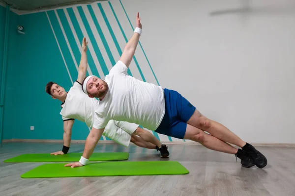 Fat man werkt hard in de fitnessruimte met zijn personal trainer doet oefeningen op de mat op de grens van de mogelijkheden — Stockfoto