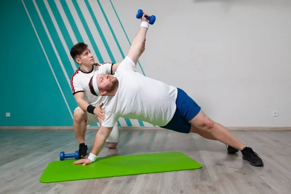 Fat man is working hard in the fitness gym with his personal trainer doing exercises with dumbbells at the limit of possibilities.